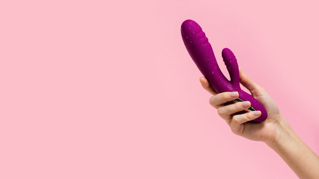 The girl's hand holds a purple vibrator covered with drops on a pink background. Vibrator for female masturbation. Dildo for vaginal and clitoris stimulation. Image for a sex shop. Sex toys for adult