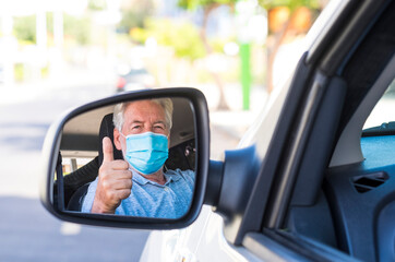 Fototapeta na wymiar Attractive smiling white-haired senior man sitting in the car wearing surgical mask while looking in the rearview mirror making the ok sign with his hand