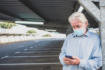 A senior man wearing medical mask due to coronavirus standing under the metal structure of a deserted parking looking at his smart phone. Nobody else