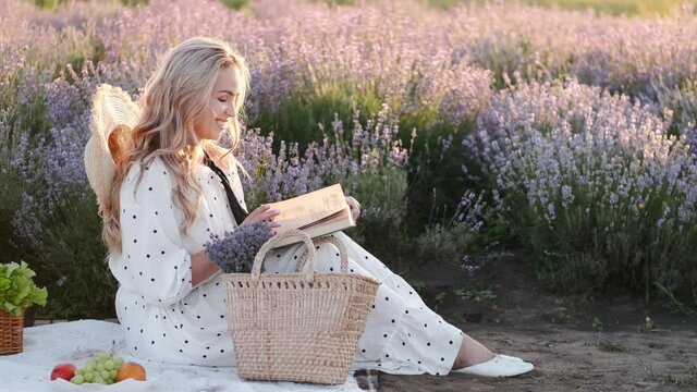 A gorgeous woman in a dress and a straw is sitting in the field and reading a book on sunset. The lady is on a picnic relaxing and taking tie for herself.