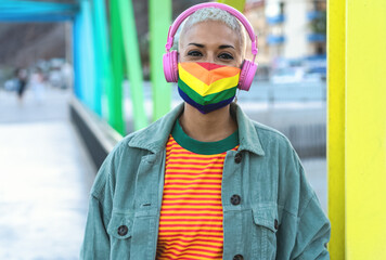 Young woman wearing gay pride mask while listening to music with headphones outdoor - Gender...