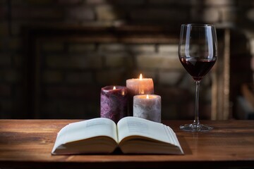 Glass of red wine, open book with candles in the background on wooden table.