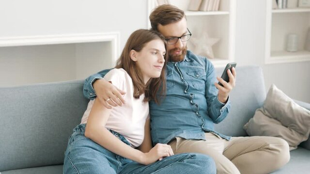 Carefree young adult couple enjoying relaxing using modern smartphone technology. Man and woman making video call playing mobile games, watching video, taking selfie sitting on a sofa together at home