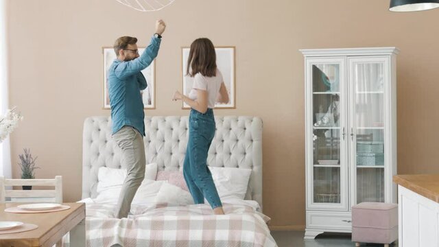 Happy funny young family couple dancing jumping on bed mattress, active carefree husband and wife having fun laughing enjoying honeymoon party together in morning in cozy bedroom interior at home
