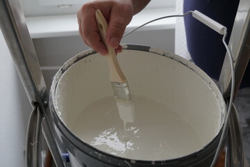 A man holding a paintbrush and dipping it in the bucket of white paint in order to paint the wall in his home. He is making improvements in his home during coronavirus lockdown. 