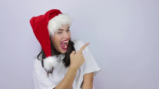 Happy young beautiful woman wearing Santa Claus hat over isolated white background pointing at copy space, showing mock workspace for advertising, empty place for text or image, promotional content.
