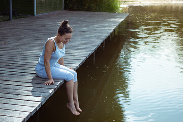 Young woman sitting on a wooden jetty dangling her legs above the water and looking down. Meditation, yoga in nature.