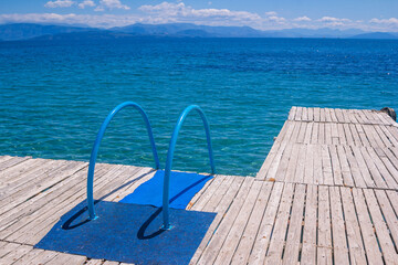 Fototapeta na wymiar View of wooden pier with blue bathing ladder. Summer landscape with turquoise calm sea water, mountains on the horizon. Corfu Island, Greece.