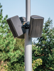 Black speakers suspended from a metal pole with blue sky as a background. Outdoor speakers for music .