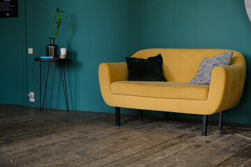 Against the blue wall is a beautiful yellow sofa with decorative pillows on four wooden legs. Part of the interior. A small table on which there is a vase with a flower.