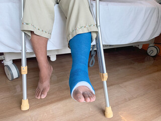 Disabled Injured Person With Sprained or Broken Ankle or Foot holding crutches siting on bed in...
