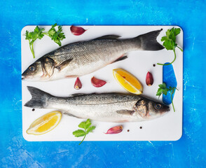 Two raw seabass with spices and lemon on white cutting board