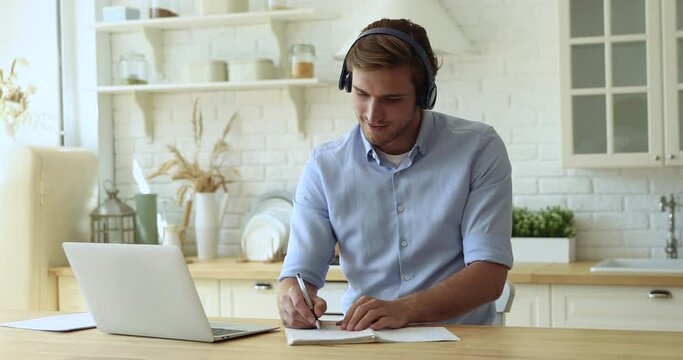 Handsome man sit at kitchen dining table learn language with e teacher wear headphones, listen tutor, writes on copybook improve english for future career advance using video conference app and laptop