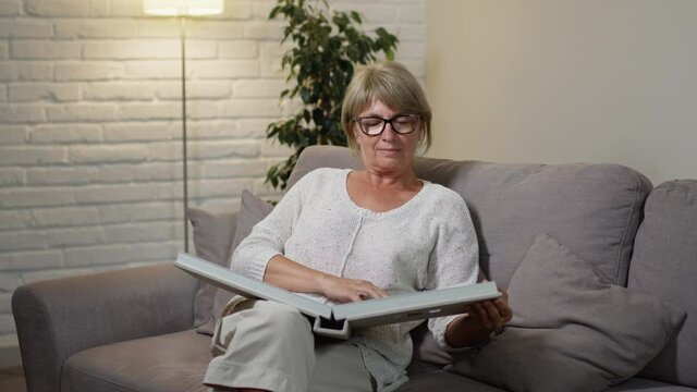 Beautiful Middle Aged Woman Sitting On The Couch In Her Apartment. Nice Blonde With Glasses In Cozy Apartment. She Holds Photo Album On His Lap And Looks At It.