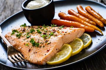 Fried salmon fillet with carrots, lemon and cream sauce served on black plate on wooden table
