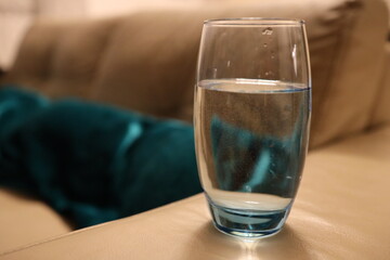 Ein trübes Glas mit Wasser aus der Nähe fotografiert. im Hintergrund ist eine braune Couch mit einem blauen Polster zu sehen.