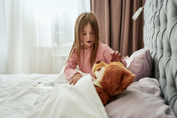 A little girl taking care of her teddybears lying next to them on a big bed covering them with a...