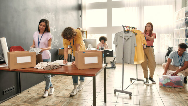 Full length shot of young volunteers packing, sorting goods, putting clothes on clothing rack for needy people, Diverse team working together on donation project indoors