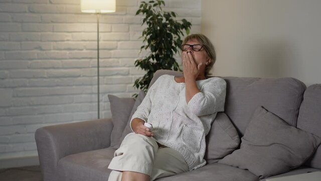 Beautiful Middle Aged Woman Sitting On The Couch In Her Apartment. She Holds The Remote From The TV And Switches Channels. Woman Is Tired. She Yawns.
