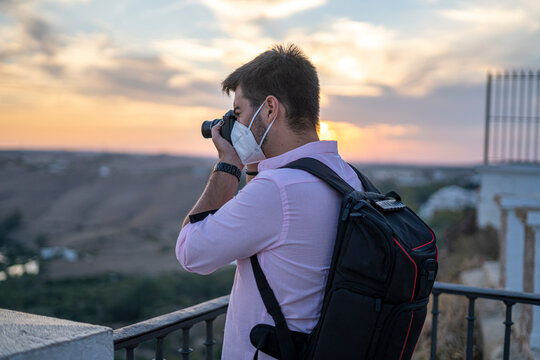 Fototapeta Hombre joven con mascarilla de viaje haciendo fotos al atardecer en un mirador del pueblo que visita