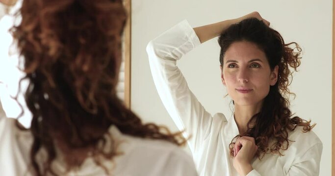 Close up attractive 35s woman in white bath robe standing in bathroom reflected in mirror touch curly brown hair looks pensive thinks about color or hairstyle change, morning routine, haircare concept