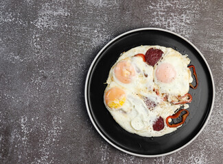 Fried eggs with pepper and sausage on on a round plate on a dark background. Top view, flat lay