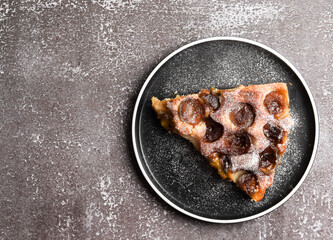 Slice of rustic plum cake on a round plate on a dark background. Top view, flat lay