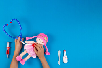 Kid hands play pretend doctor game. Toy stethoscope, pink doll and medicine tools on light blue...