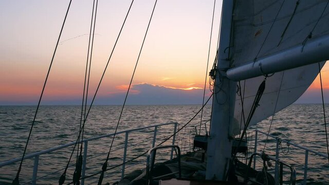 A sailing yacht is going towards the setting sun. Beautiful sunset sky ahead. The sails are full of wind.	
