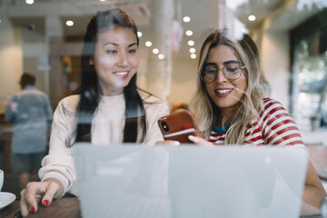 Cheerful multiracial female students spending time together searching information online for project,young women watching vlog on smartphone together enjoying free time on break and blogging