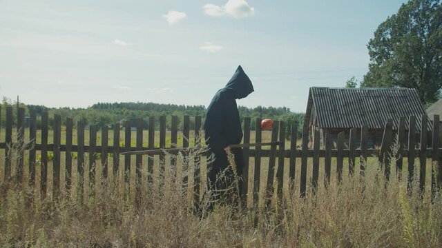 Scary creepy grim reaper with rustic scythe coming to collect lost souls in village on halloween holiday. Dark angel of death in shroud standing near wooden fence with halloween pumpkin stuck in it.