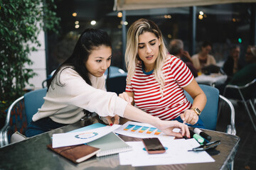 Successful hipster girls talking during collaborative meeting for checking informative documents in street cafe,multiracial female planning organisation while doing university homework and course work