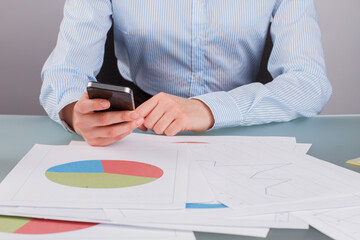 Female hands using smartphone while working at desk in office. Business woman analyzing graph data and checking mail on smartphone.