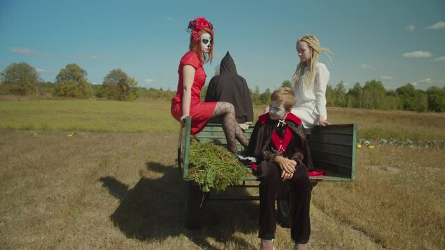 Dark angel of death in black shroud driving mini tractor with people dressed in halloween costumes and wearing makeup, seated in trailer while grim reaper transporting souls of deceased to afterlife.