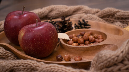Autumn still life with apples and nuts. Autumn background with apples on a warm knitted scarf, a wooden plate, autumn leaves, hazelnuts and cones.