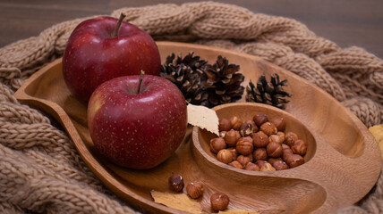 Autumn still life with apples and nuts. Autumn background with apples on a warm knitted scarf, a wooden plate, autumn leaves, hazelnuts and cones.