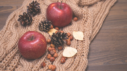 Autumn still life with apples and nuts. Autumn background with apples on a warm knitted scarf, a wooden plate, autumn leaves, hazelnuts and cones.