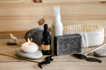 Fototapeta na wymiar Above view set of various man spa products things. Natural wood disc background with flat sea stones. Gray clay soap bar, beard oil, moisturizing cream, candle burning. Funny mustache shape figurines.