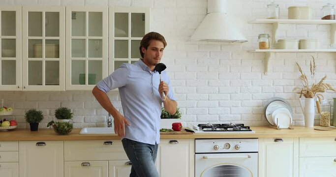 Handsome happy man singing dancing alone in cozy kitchen holds ladle like microphone. Lively active guy imagining himself like a star, enjoy weekend distracted from food preparation having fun at home
