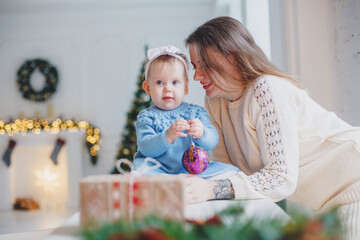 Mother with daughter in knitted clothes in a bright room at a table. Christmas atmosphere - fireplace, gifts, bokeh.