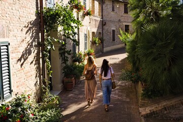 A couple of girls is walking in an italian medieval village with plants and flowers on the street (Corinaldo, Marche, Italy, Europe)