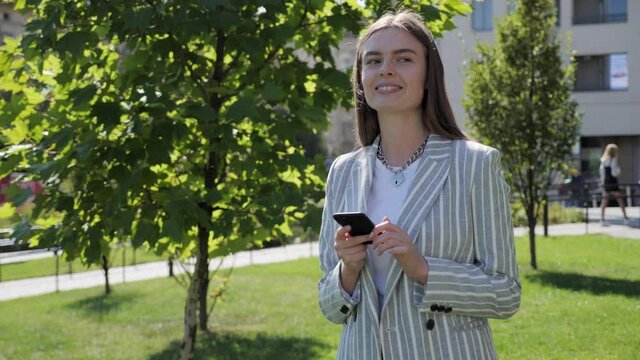 Successful happy businesswoman in light grey suit is texting messages on her modern smartphone outdoors. Young girl having good mood, looking satisfied. Successful Lifestyle.