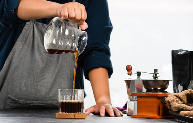 Women barist is pouring hot coffee from the coffee pot into the glass coffee on the wooden table in the morning. Woman to make a drip brewing and drinking hot black coffee.