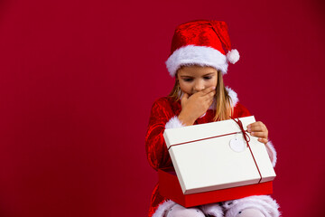 Portrait of a thinking young girl with a open gift box isolated over red background, dressed in santa hat and costume
