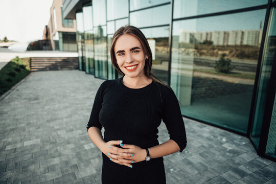 Portrait of young confident smiling businesswoman standing outside near glass office building, dressed in black dress, wear hand watch and looking at the camera. Image with copy space.