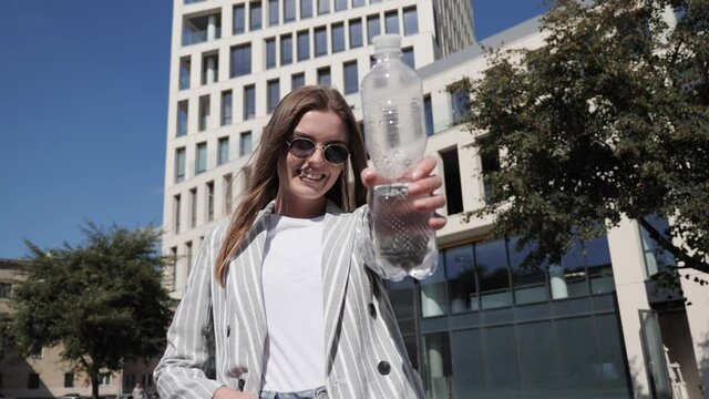 Beautiful girl in a light jacket and sunglasses drinks water and then smiles on the background of the business center.