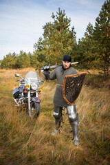 A medieval knight in chainmail with a shield and a sword in his hands stands against the backdrop of a motorcycle and a forest.