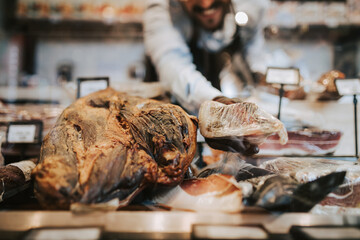 Handsome middle age male seller working at supermarket or grocery store and selling cured meat products and cheeses.