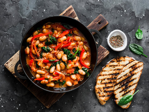 Greek style tomato sauce, spinach, paprika, beans stew in a cast iron pan on a rustic board on a dark background, top view. Simple comfort food