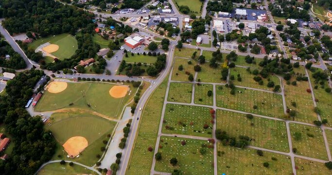 We fly over baseball fields for recreation and Pine Hill cemetery with monuments and headstones visible. Burlington, NC. Beside the baseball fields is an apartment complex.